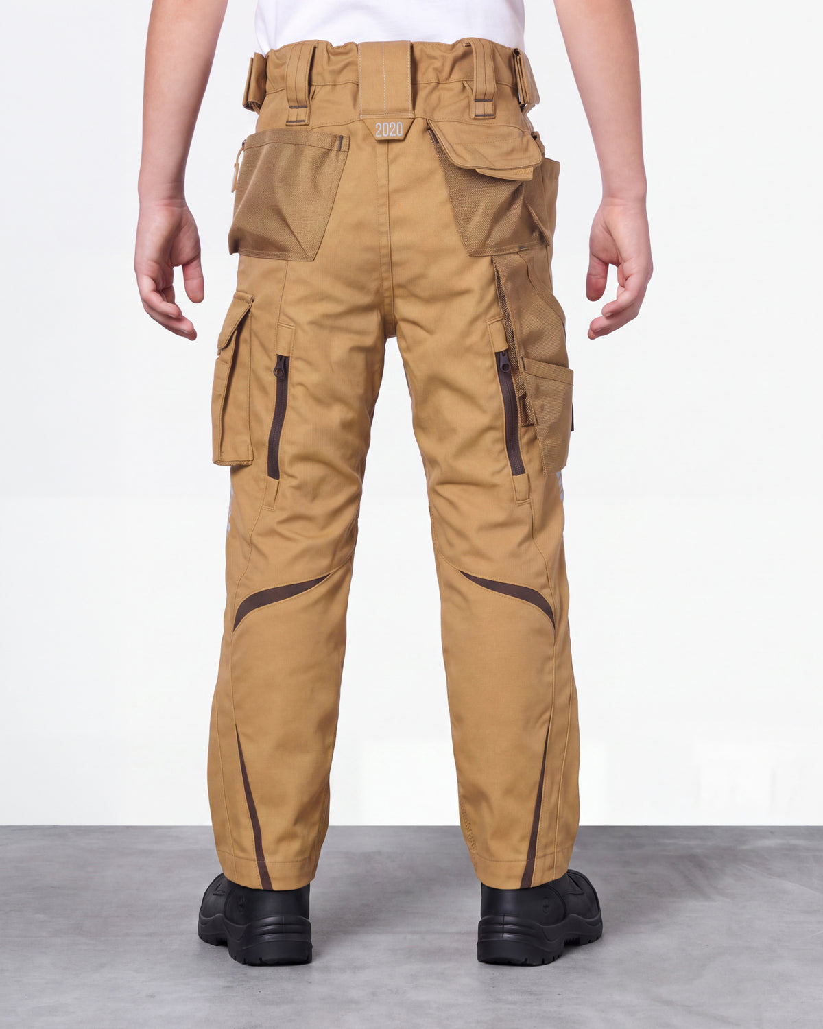 A person shown from the back wearing tan cargo work pants with multiple flap pockets, utility pockets, and dark contrast piping, paired with black shoes against a plain light gray studio background.