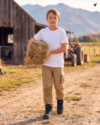 A young boy stands on a dirt path at a farm holding a round bale of hay. He is wearing a white t-shirt, tan cargo pants, and black boots, with a wooden barn, tractor, and mountains in the background.