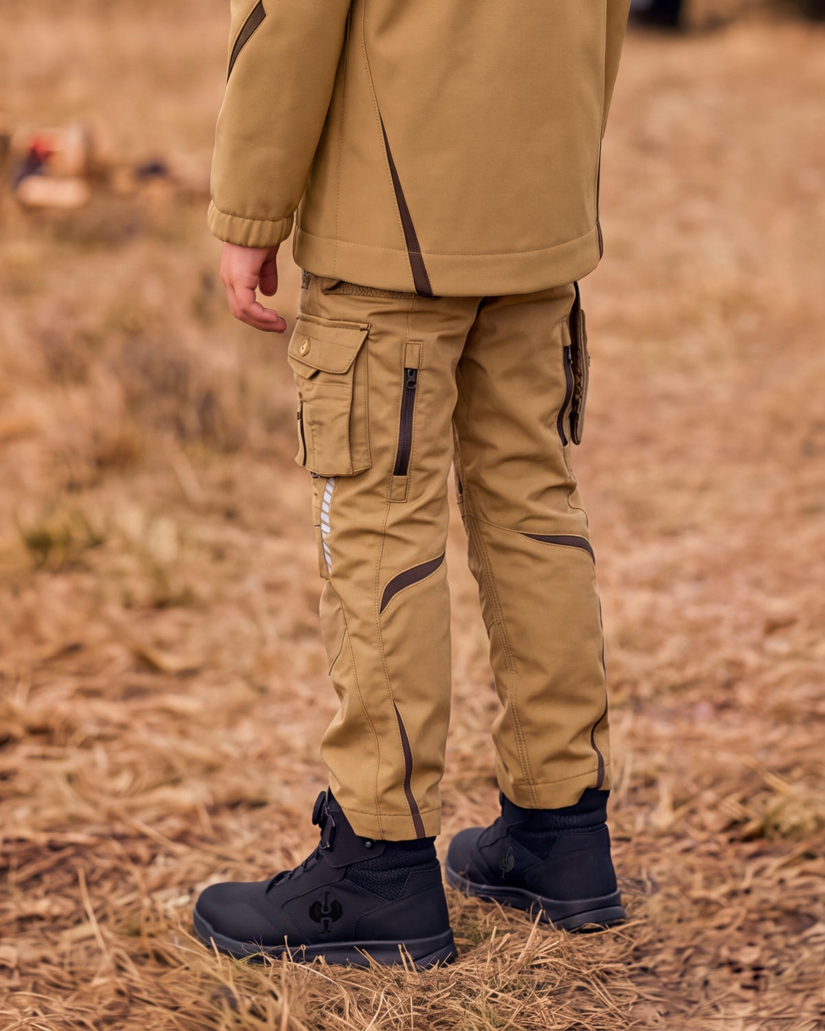 A person outdoors wearing tan cargo pants with dark contrast stitching and zippers, a matching tan jacket, and black boots on dry grass.