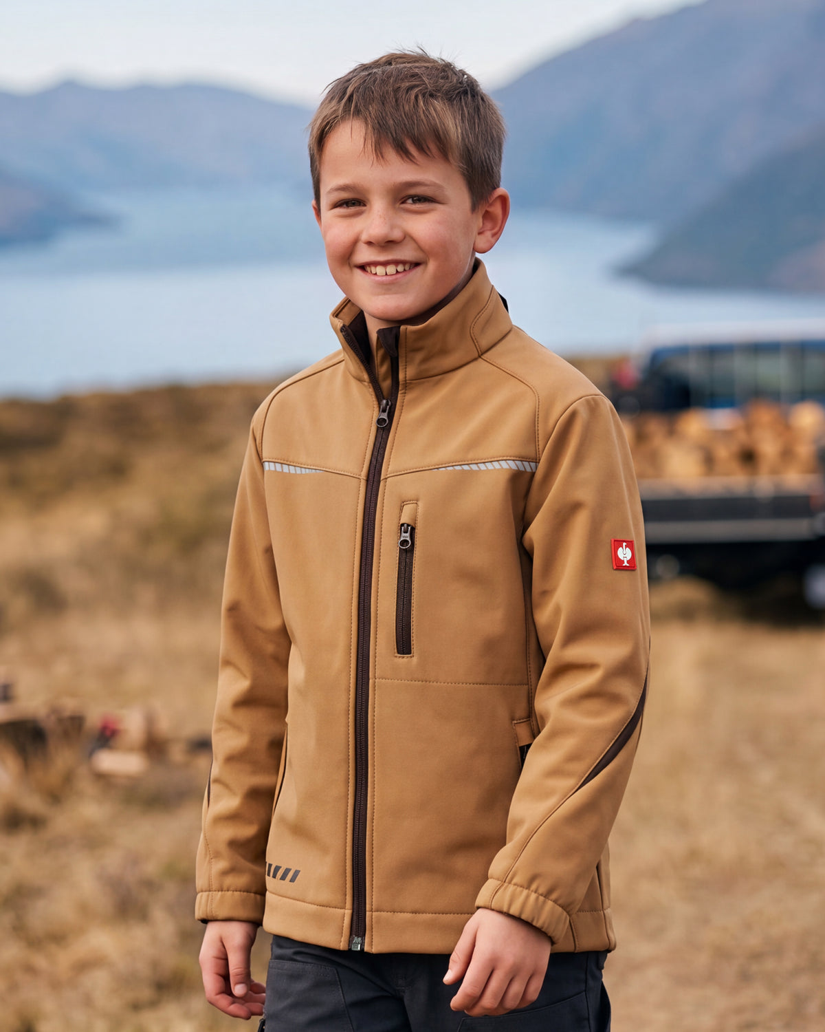 A smiling young boy wearing a tan zip-up jacket with reflective chest stripes and a red Swiss cross logo on the sleeve, standing outdoors in front of a blurred mountain and lake background.