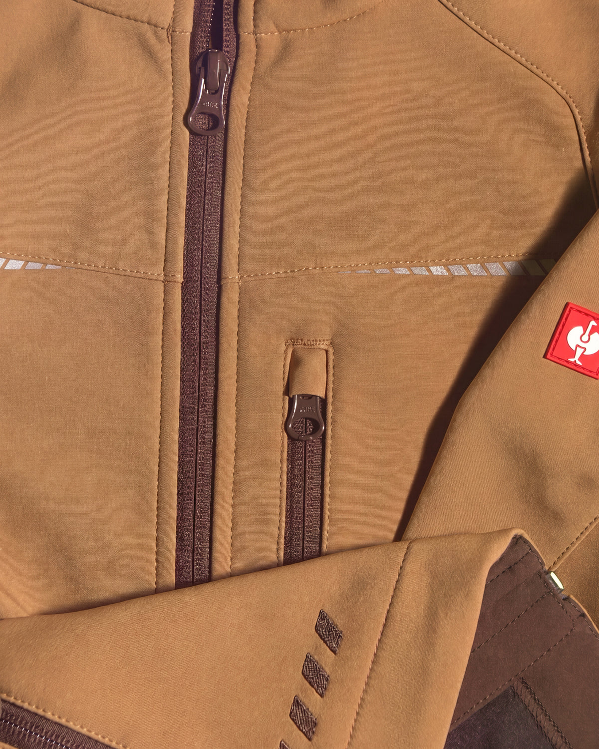 Close-up of a tan technical jacket with dark brown zippers, a small red square logo patch on the sleeve, and reflective white stripe details.