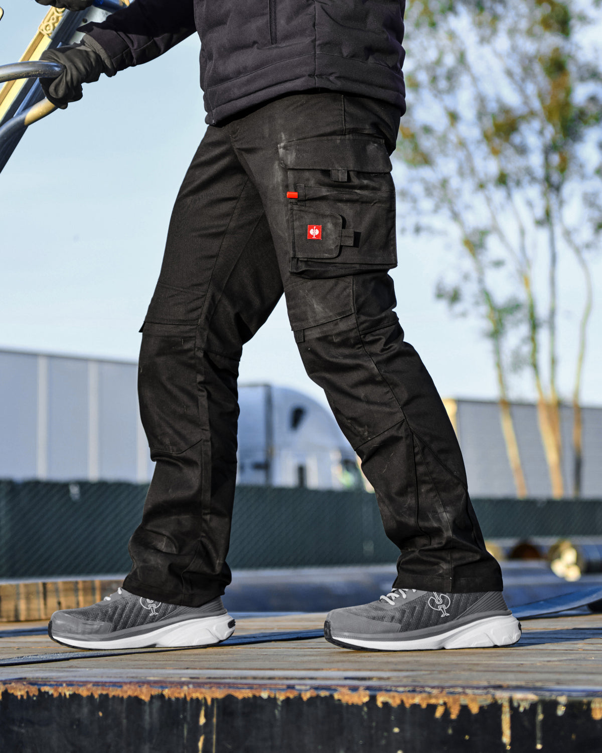 Black cargo work pants worn by a model outdoors, featuring multiple utility pockets and a small red logo patch on the thigh, paired with gray sneakers.