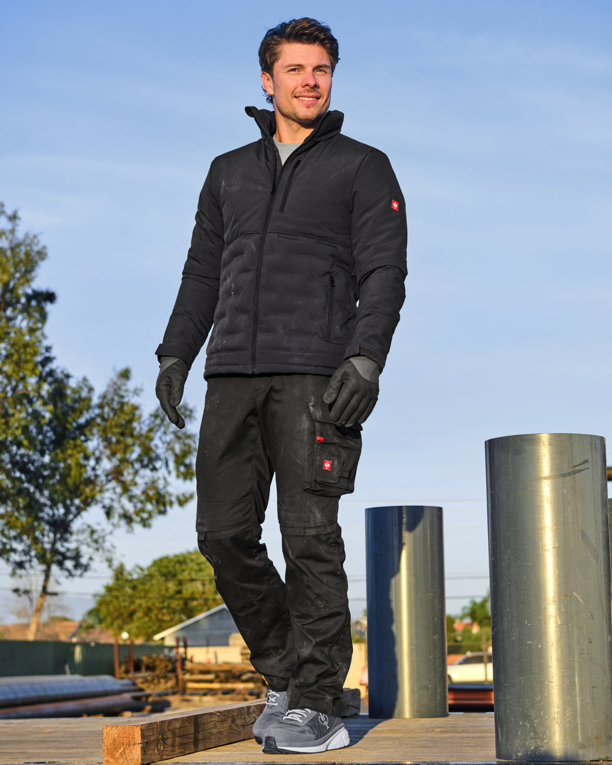 A man wearing a black insulated jacket, black cargo pants, black gloves, and gray sneakers, standing outdoors on a wooden deck next to metal columns under a blue sky.