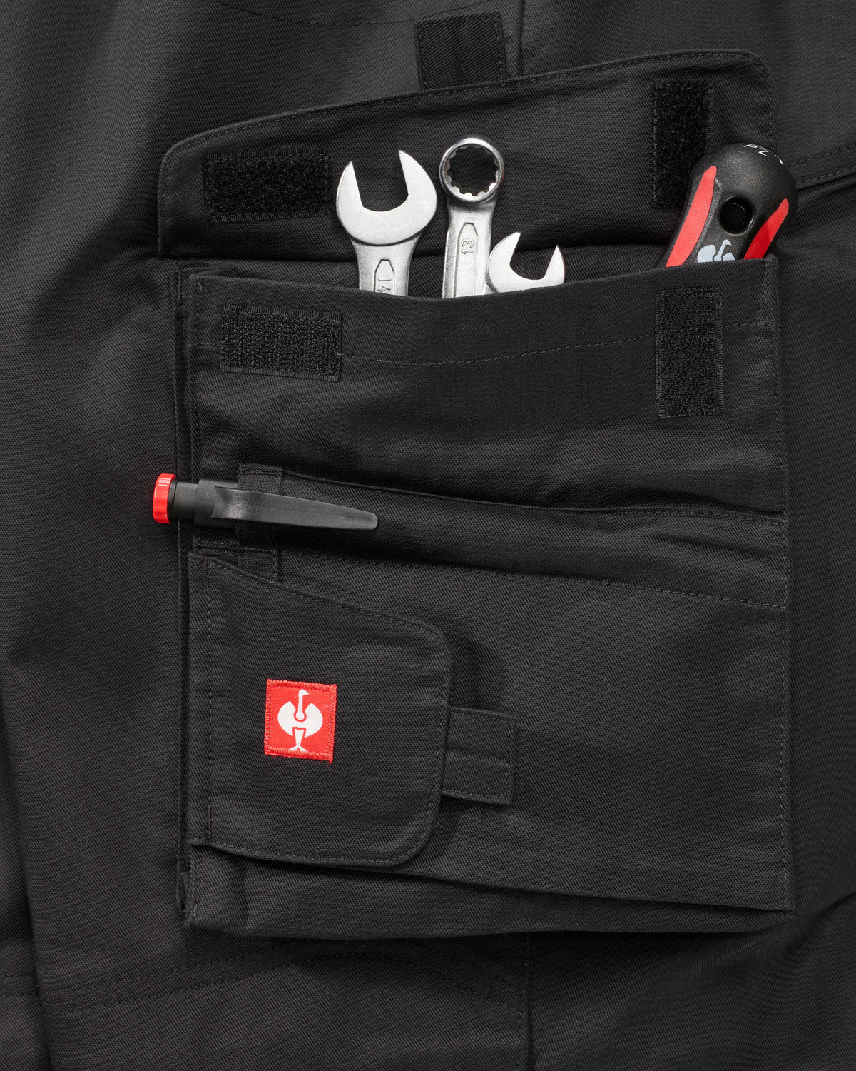 Close-up of a black workwear utility pocket with tools inserted, including wrenches and a pen, featuring a small red square logo patch on the front flap.