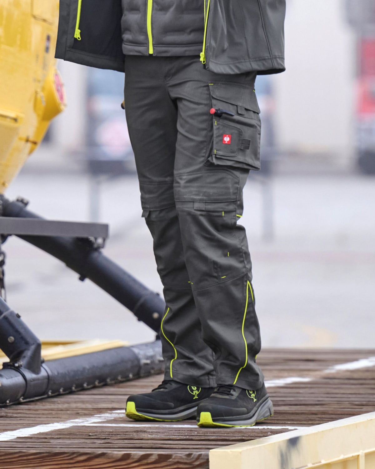 A person wearing dark gray technical cargo pants with bright neon yellow-green piping and zippers, paired with black and neon yellow-green work boots, standing on a wooden platform.