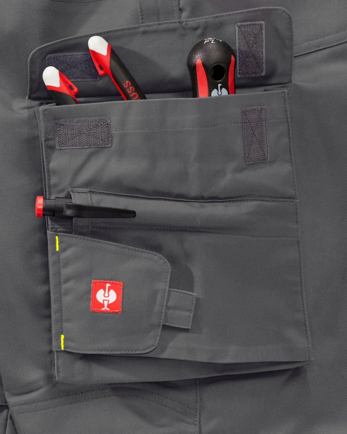 Close-up of a dark gray tool pouch on workwear pants, holding hand tools, with a red square logo patch on the front pocket.