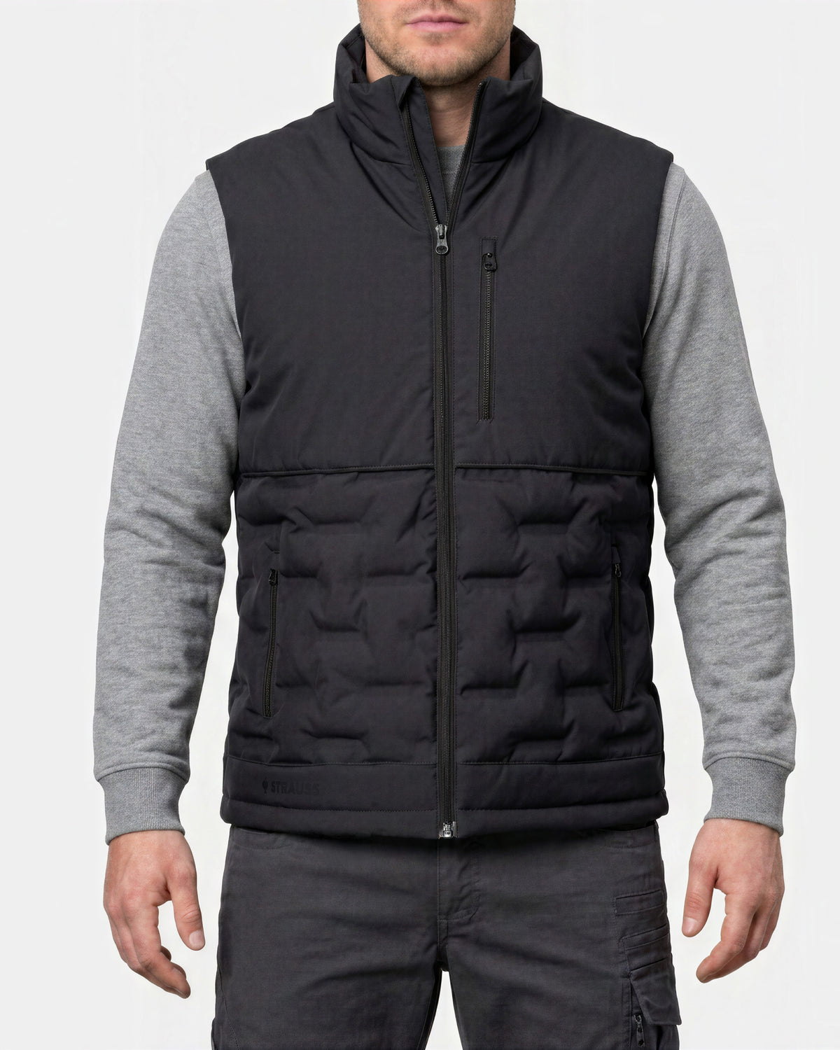 A man wearing a black quilted zip-up puffer vest over a gray long-sleeve shirt and dark gray pants, shown from the mouth down against a light gray studio background.