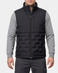 A man wearing a black quilted zip-up puffer vest over a gray long-sleeve shirt and dark gray pants, shown from the mouth down against a light gray studio background.