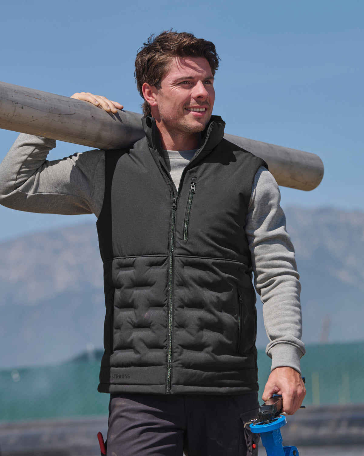 A man wearing a black insulated zip-front vest over a gray long-sleeve shirt, carrying a large wooden log on his shoulder outdoors with mountains and blue sky in the background.