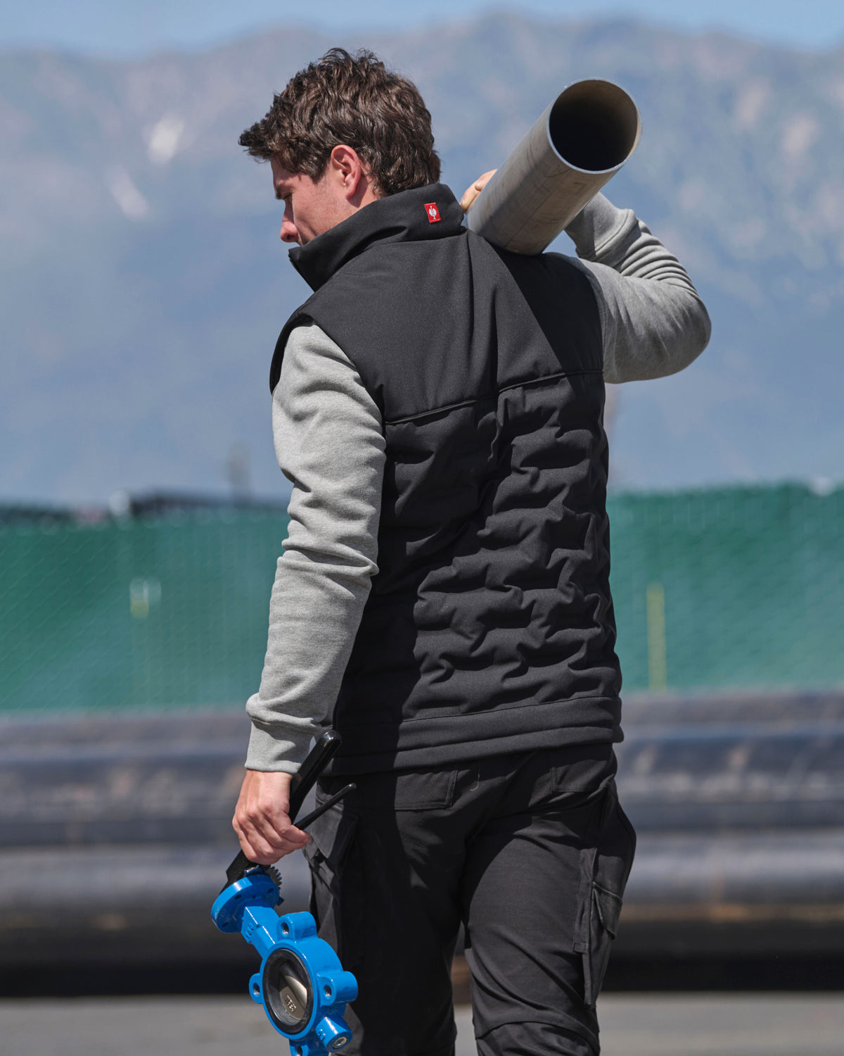 A man wearing a black quilted vest over a gray long-sleeve shirt walks outdoors carrying a large metal pipe on his shoulder and a blue industrial valve in his hand, with mountains and a green fence in the background.