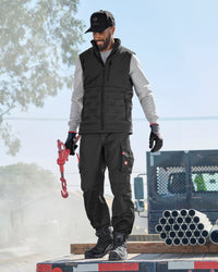 A man wearing a black insulated work vest over a light gray long-sleeve shirt, black work pants, black gloves, and a black cap stands on a truck bed holding a red hook. The background shows an outdoor construction site with pipes and a vehicle.