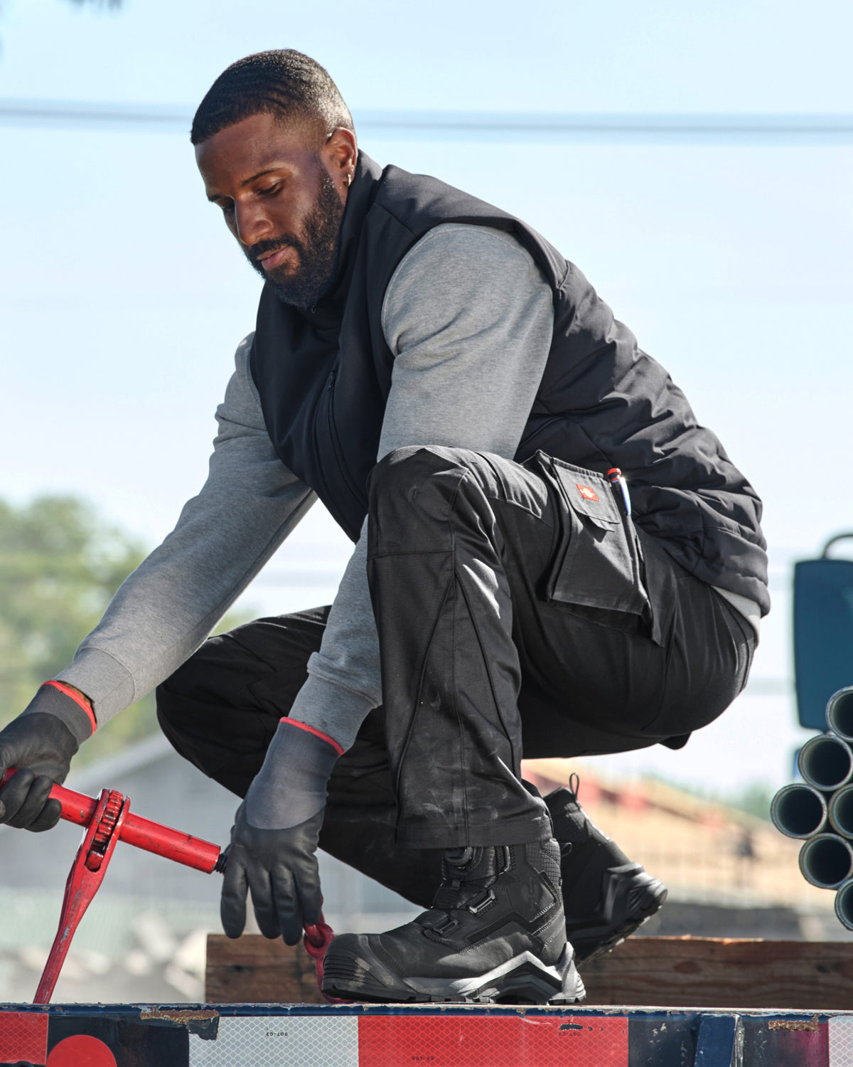 A man in workwear crouches on a truck bed, wearing a black quilted vest over a gray long-sleeve shirt, black work pants, black gloves, and black work boots, while using a red hand tool.