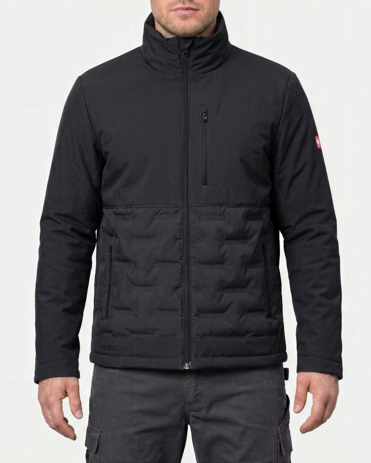 A man wearing a black technical jacket with a high collar, full zip front, and horizontal quilted paneling on the lower half, shown against a light gray studio background.