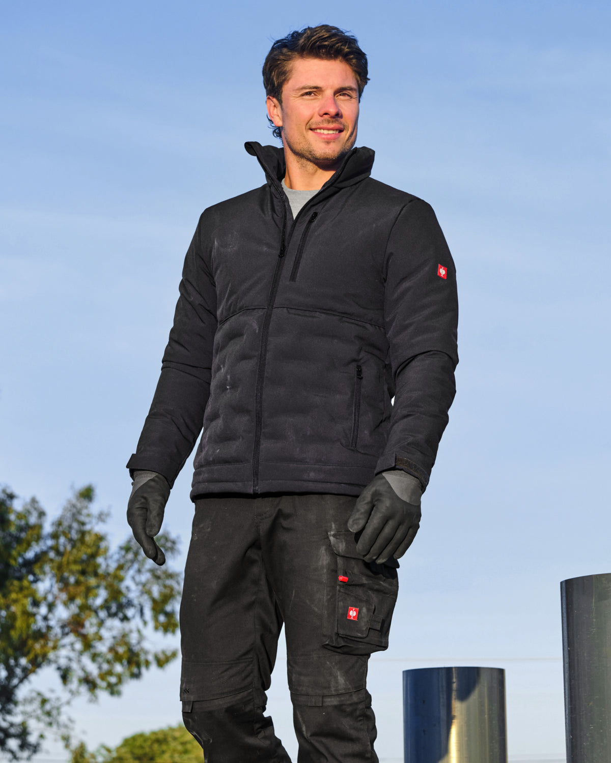 A man wearing a black technical fleece jacket, black work pants, and black gloves, standing outdoors against a blue sky.
