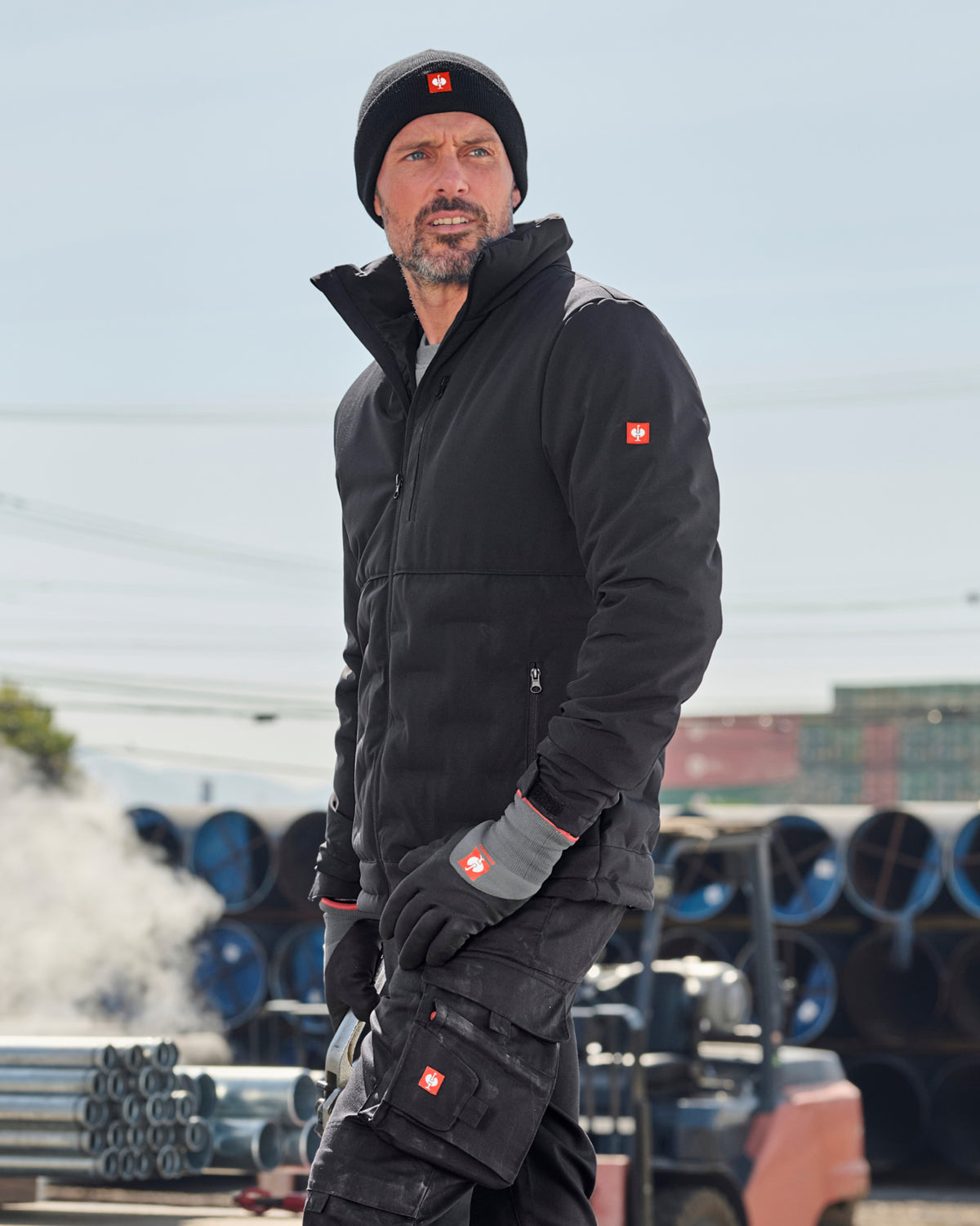 A man wearing a black technical jacket, black beanie, and black work gloves with a red Swiss cross logo, standing outdoors in front of a light blue sky and stacks of blue pipes.