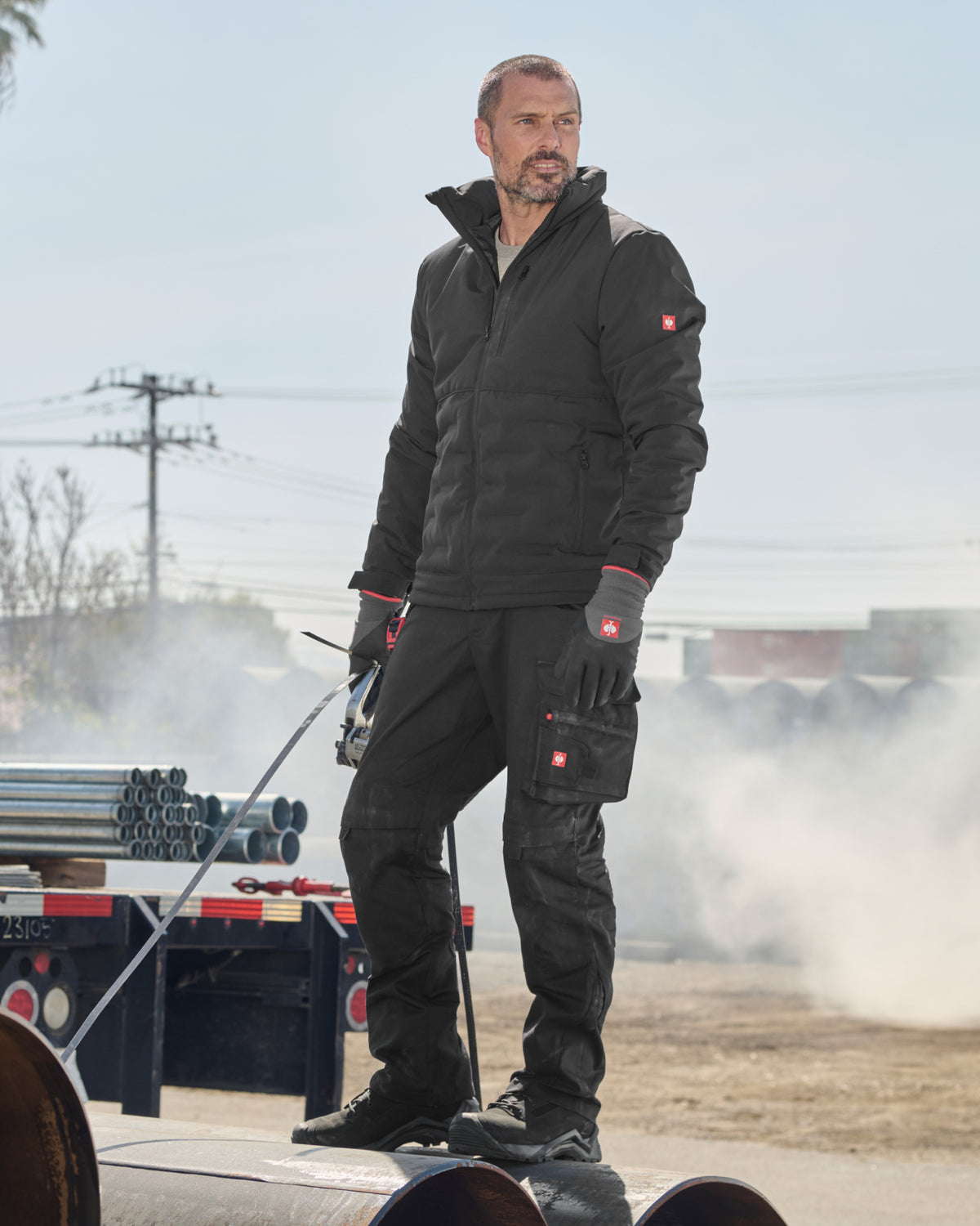 A man wearing a black insulated work jacket and matching black work pants with red logo accents, standing outdoors on a large pipe in a dusty industrial setting.