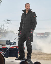 A man wearing a black insulated work jacket and matching black work pants with red logo accents, standing outdoors on a large pipe in a dusty industrial setting.