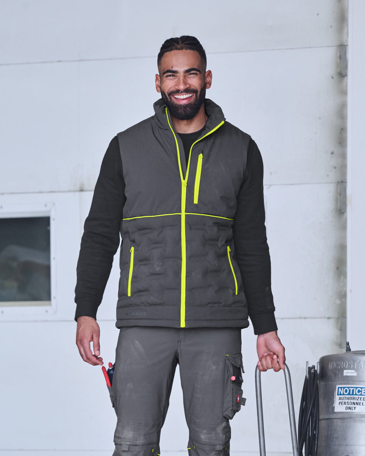 A smiling man wearing a dark gray quilted work vest with bright yellow-green piping over a black long-sleeve shirt and gray work pants, standing in front of a white industrial wall.