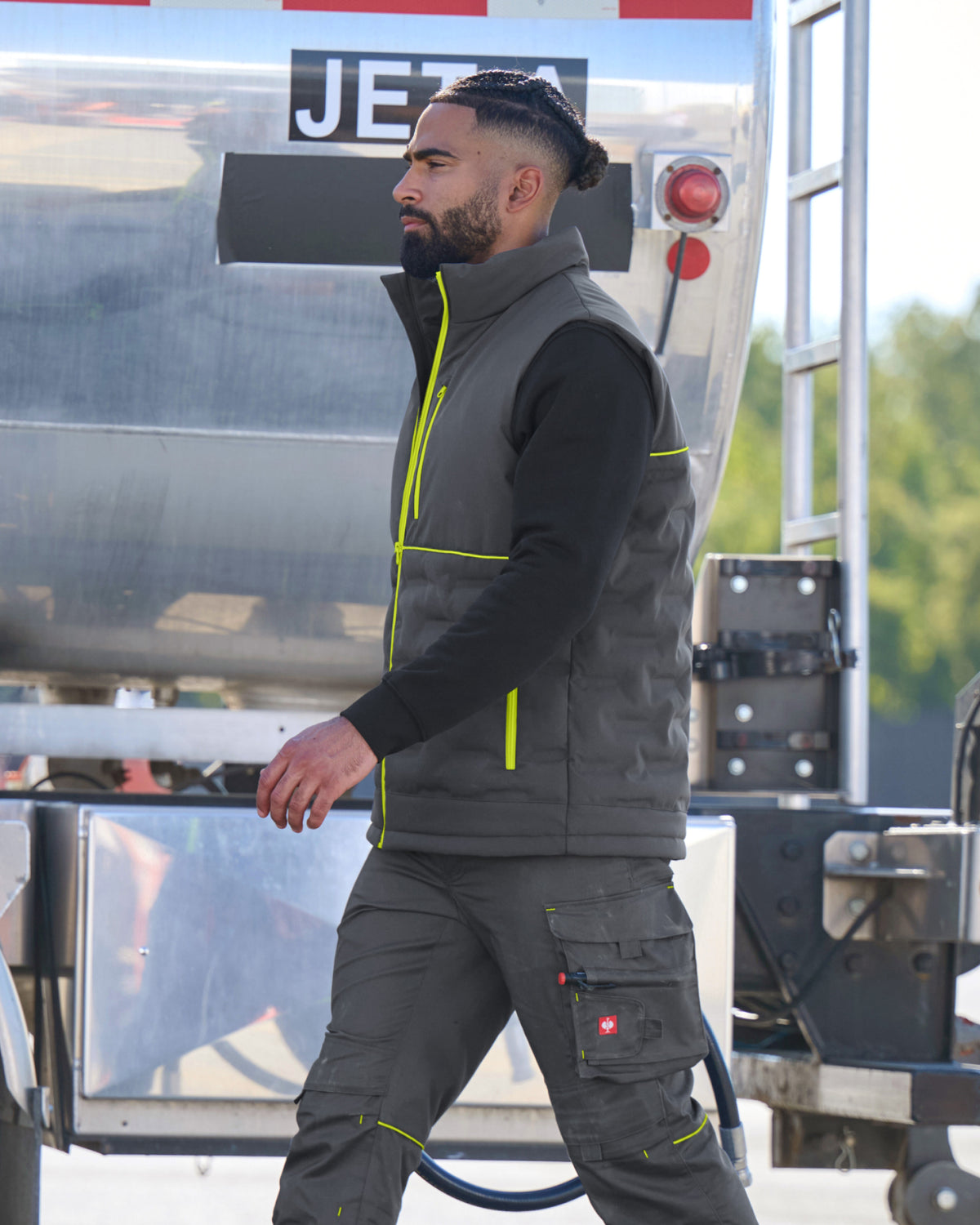 A man wearing a dark gray insulated work vest with bright yellow-green piping over a black long-sleeve shirt and matching dark gray work pants, standing in front of a large silver tanker truck.