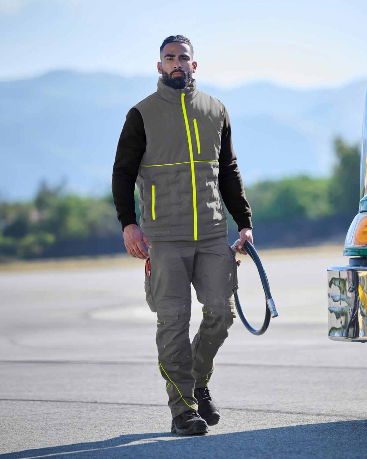 A man wearing a gray and black high-visibility work vest with neon yellow accents, matching gray work pants, and black boots, walking on an outdoor paved surface with mountains in the background.