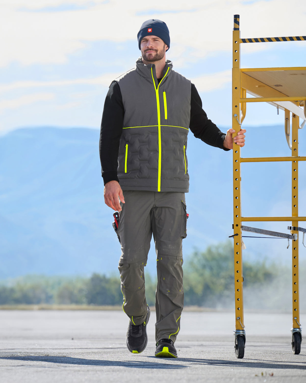 A man wearing a gray insulated work vest with bright yellow accents, matching gray work pants, and a black beanie walks on an outdoor paved surface next to a yellow scaffolding structure.