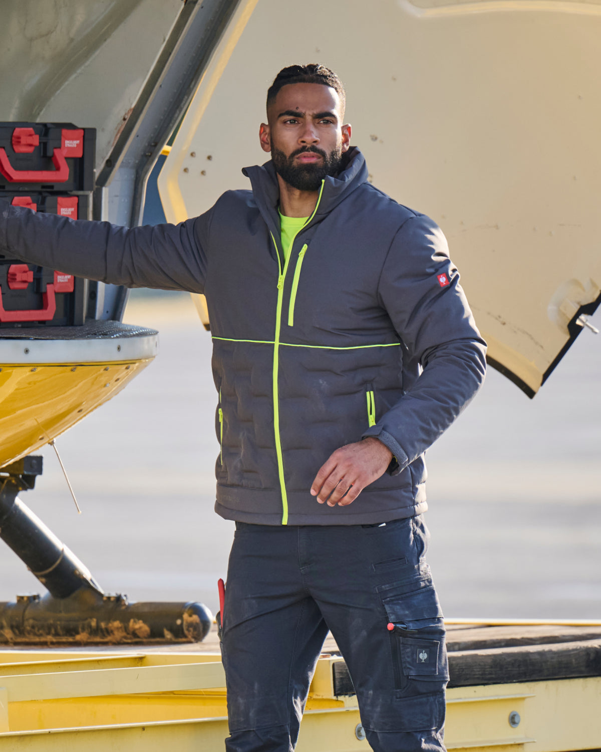 A man wearing a dark gray puffer jacket with bright neon green zippers and a matching dark cargo pants, standing next to a yellow aircraft on an airfield.