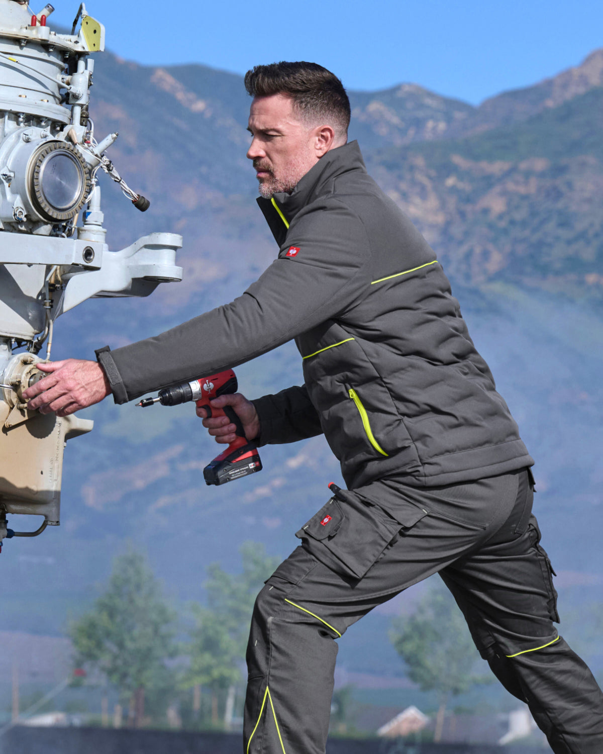 A man wearing a dark gray technical workwear set with neon yellow-green piping and a red logo patch, working outdoors next to a machine with mountains in the background.