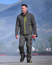 A man wearing a dark gray insulated jacket and matching dark gray work pants with neon yellow accents, walking outdoors on a paved surface with mountains in the background.