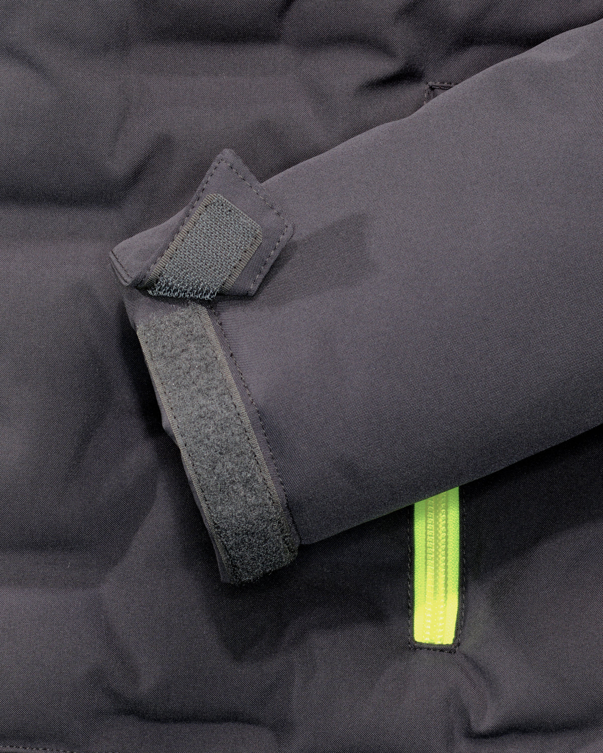 Close-up of a dark gray puffer jacket sleeve showing a hook-and-loop strap closure and a bright neon yellow zipper pull.