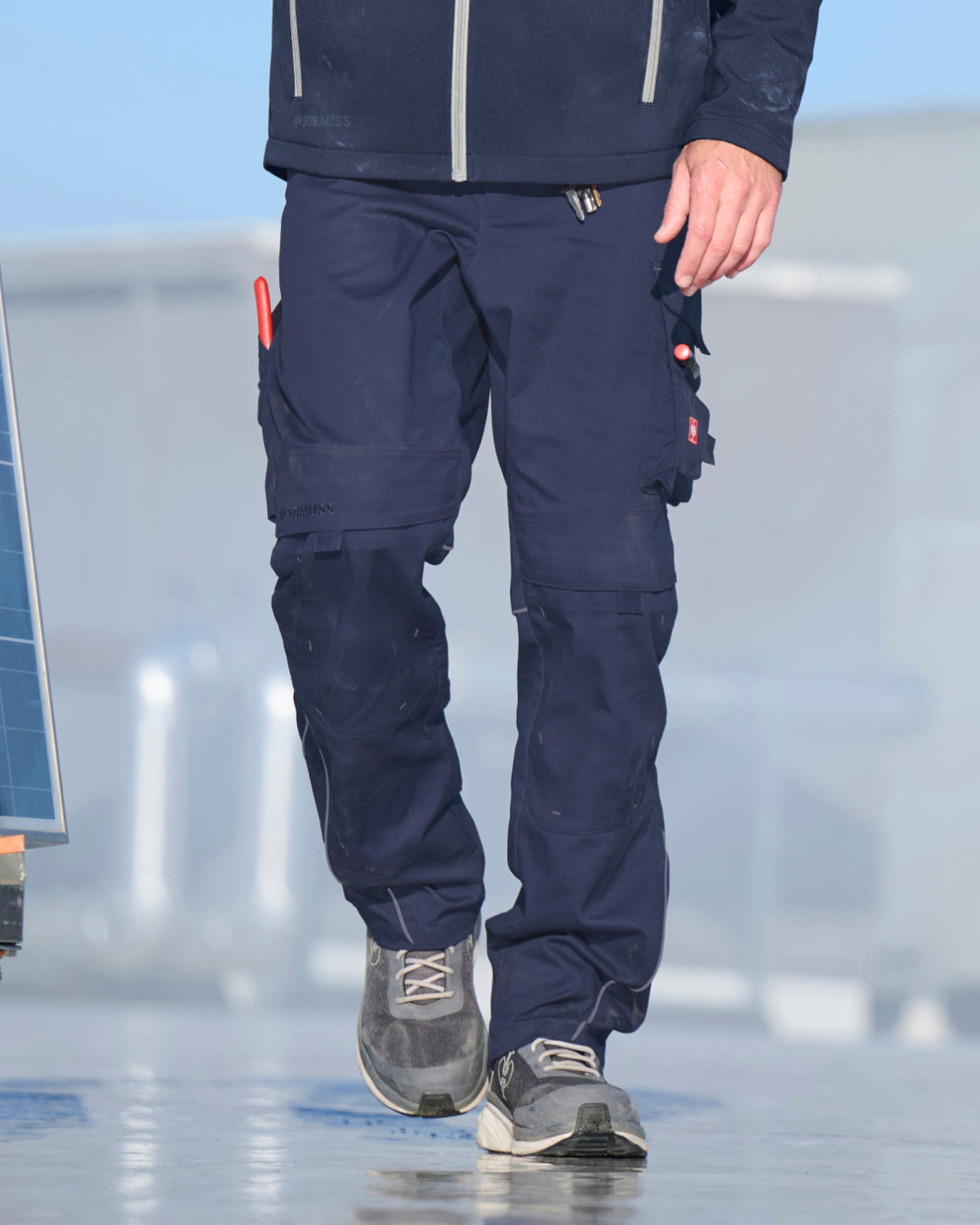 A person wearing dark navy blue work pants with multiple utility pockets and gray sneakers, shown from the waist down in an outdoor setting.