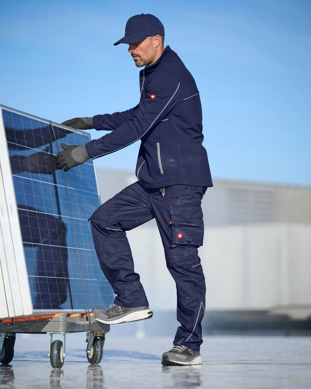 A man in a navy blue work uniform and cap pushes a solar panel on a wheeled dolly outdoors under a clear blue sky.