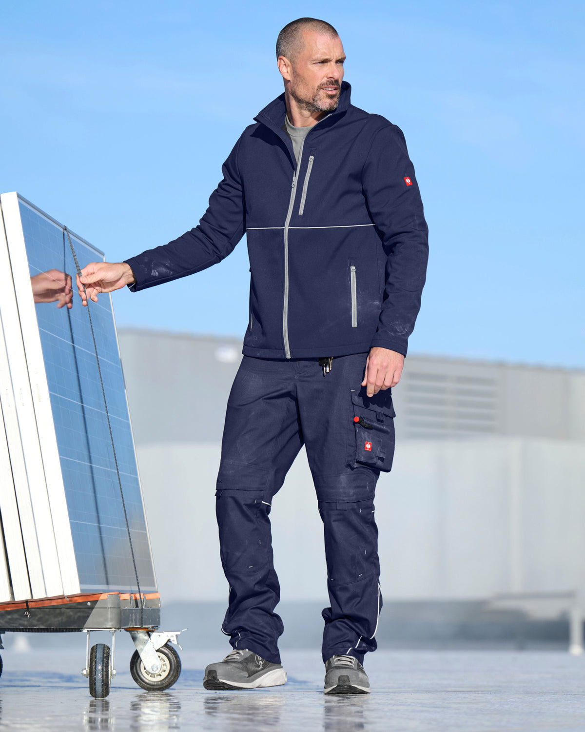 A man wearing a navy blue technical work jacket and matching cargo pants stands outdoors next to a cart holding a large glass panel, with a clear blue sky and industrial building in the background.