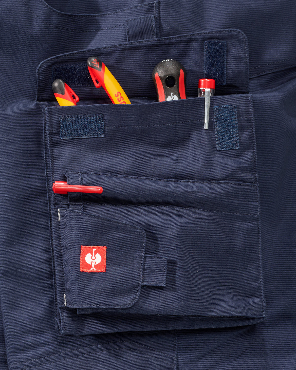 Close-up of a navy blue work pants pocket holding various hand tools, featuring a small red square patch with a white rooster logo on the front flap.