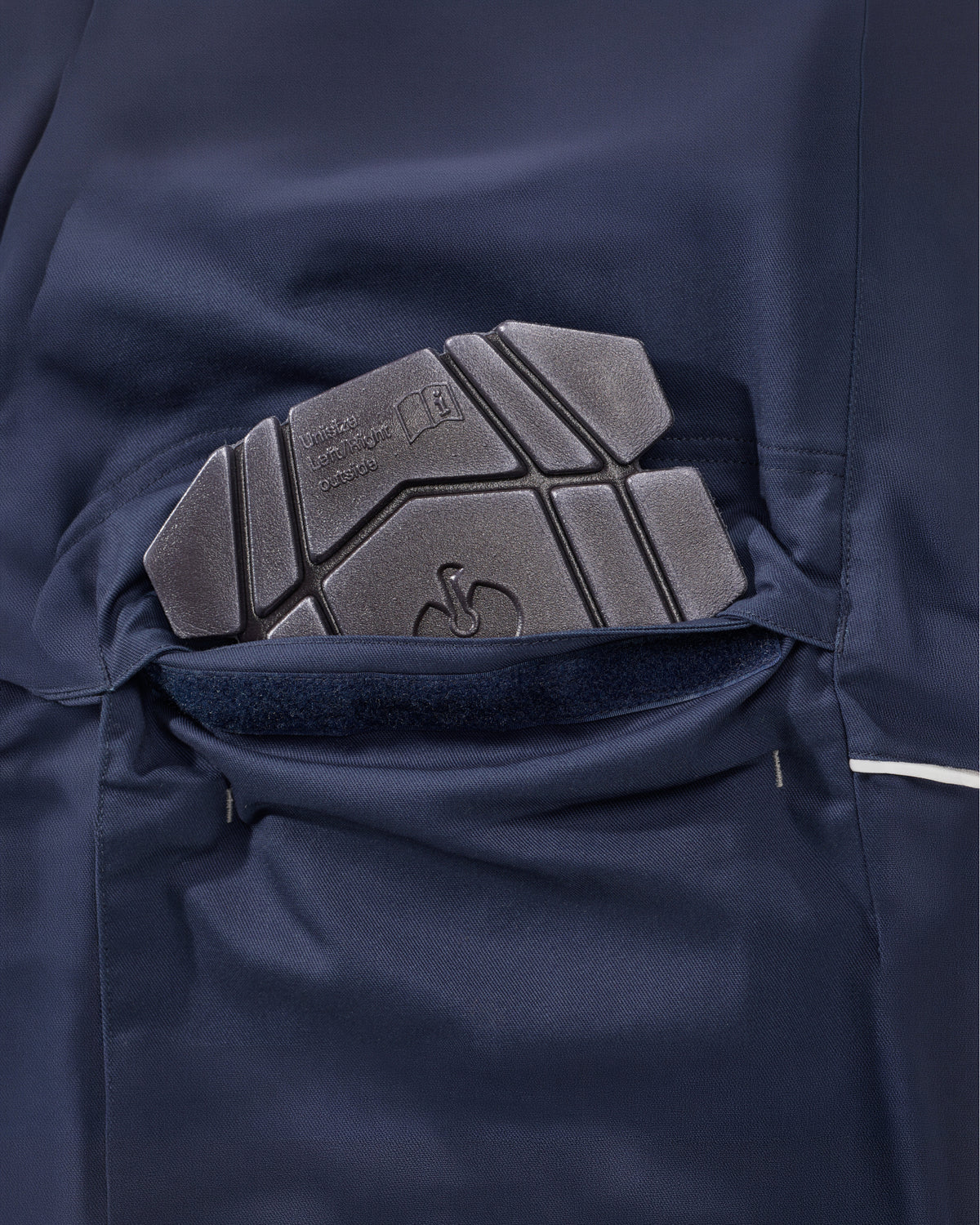 Close-up of a dark navy work pants knee pad pocket with a textured dark gray protective pad partially pulled out, showing the interior lining.