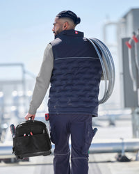 A man in a navy blue quilted work vest, navy pants, and a gray long-sleeve shirt walks away from the camera carrying a black tool bag and a coiled cable over his shoulder in an industrial outdoor setting.