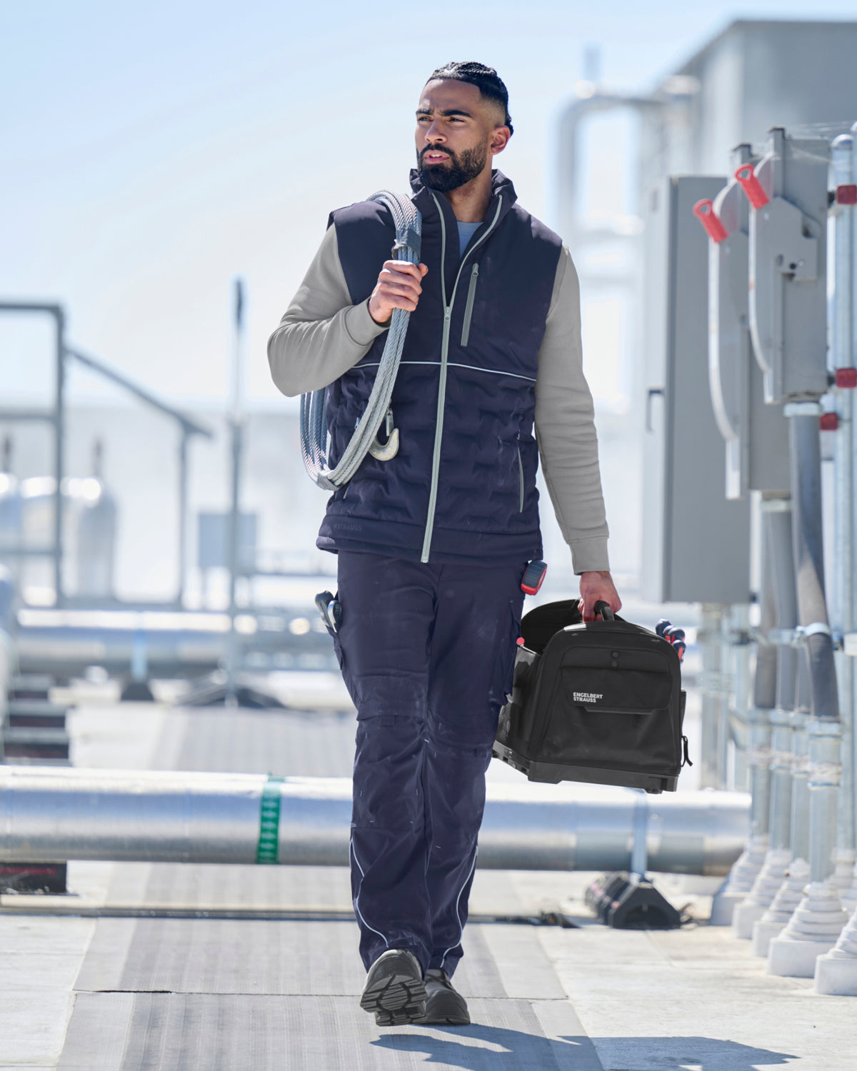 A man walks outdoors on a sunny day wearing a navy blue zippered vest over a light gray long-sleeve shirt and matching navy pants, while carrying a black bag.