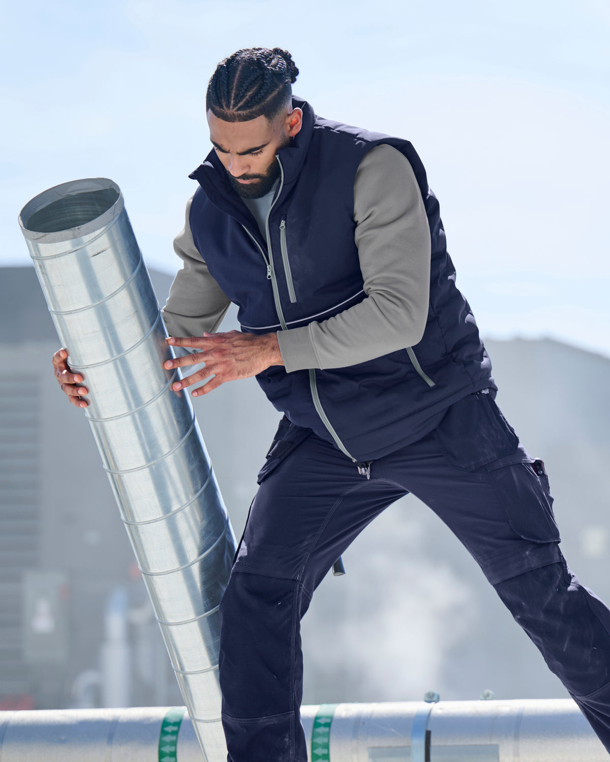 A man in a navy and gray work uniform pushes a large galvanized steel ventilation duct outdoors on a rooftop.
