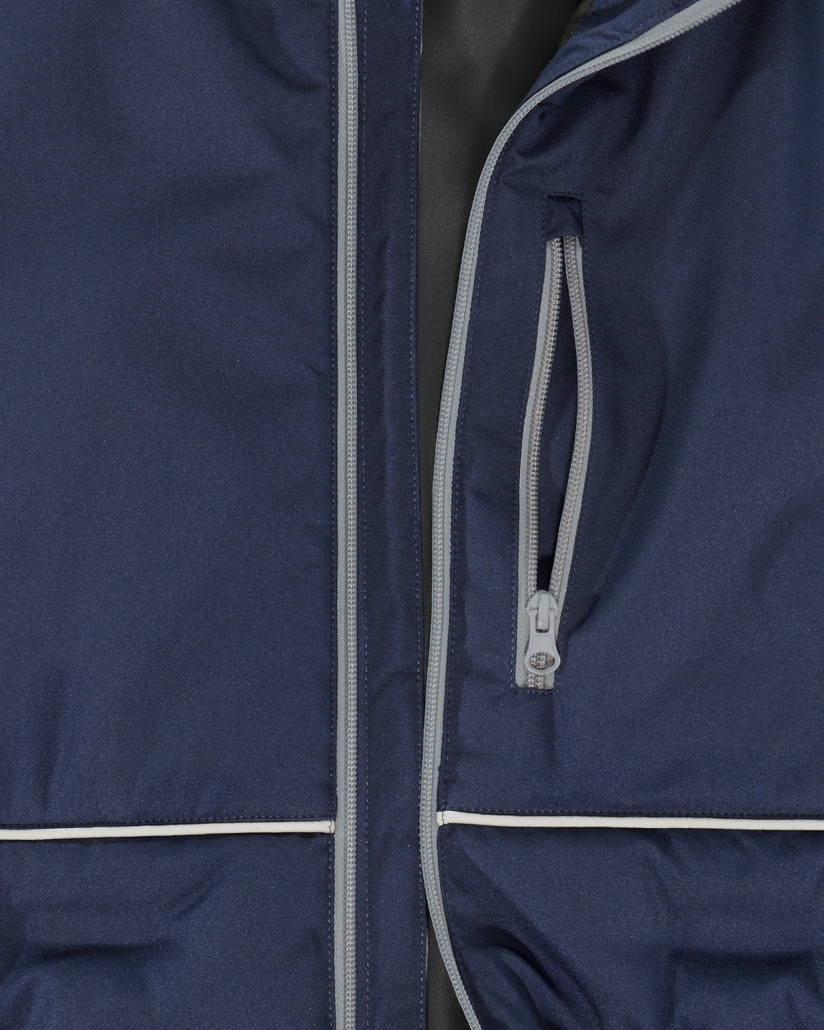 Close-up of a navy blue jacket with light gray piping and a zippered pocket, showing the fabric texture and zipper details.