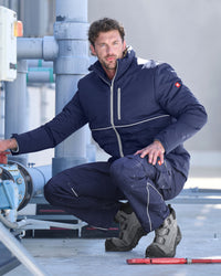 A man crouches outdoors in a navy blue technical winter jacket and matching pants with light piping, paired with gray work boots, against an industrial background of pipes and concrete.