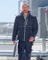 A man wearing a navy blue puffer jacket with light gray piping and matching navy pants, standing outdoors on a platform and holding a long tool.