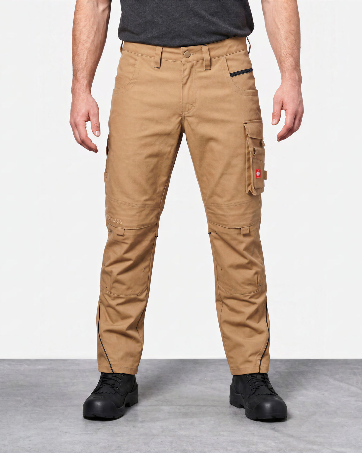 A model from the waist down wearing tan cargo work pants with black zippers and black work boots, set against a plain studio background.