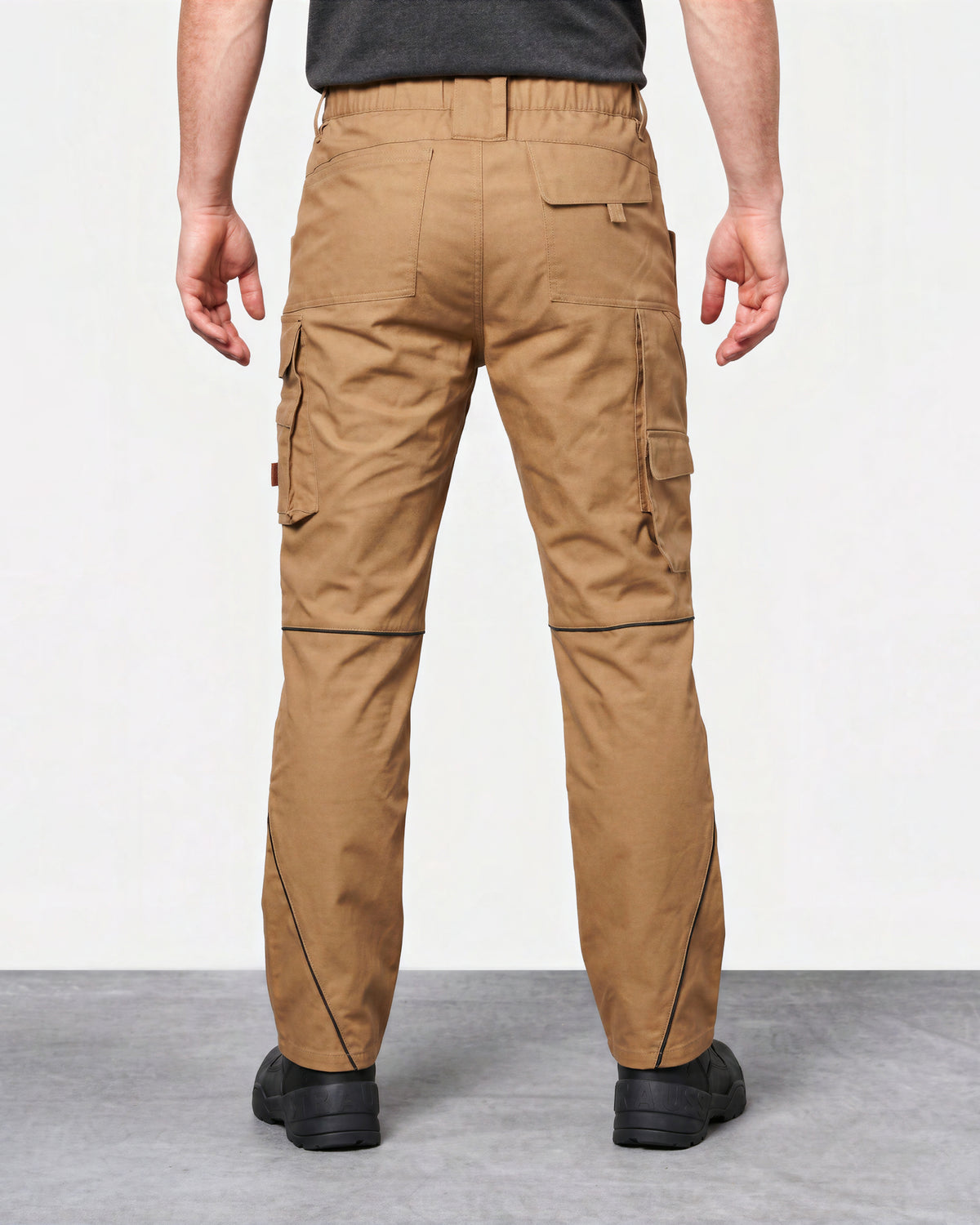 A person shown from the back wearing tan cargo work pants with black shoes, photographed in a studio setting with a light gray floor and white wall.
