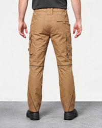 A person shown from the back wearing tan cargo work pants with black shoes, photographed in a studio setting with a light gray floor and white wall.