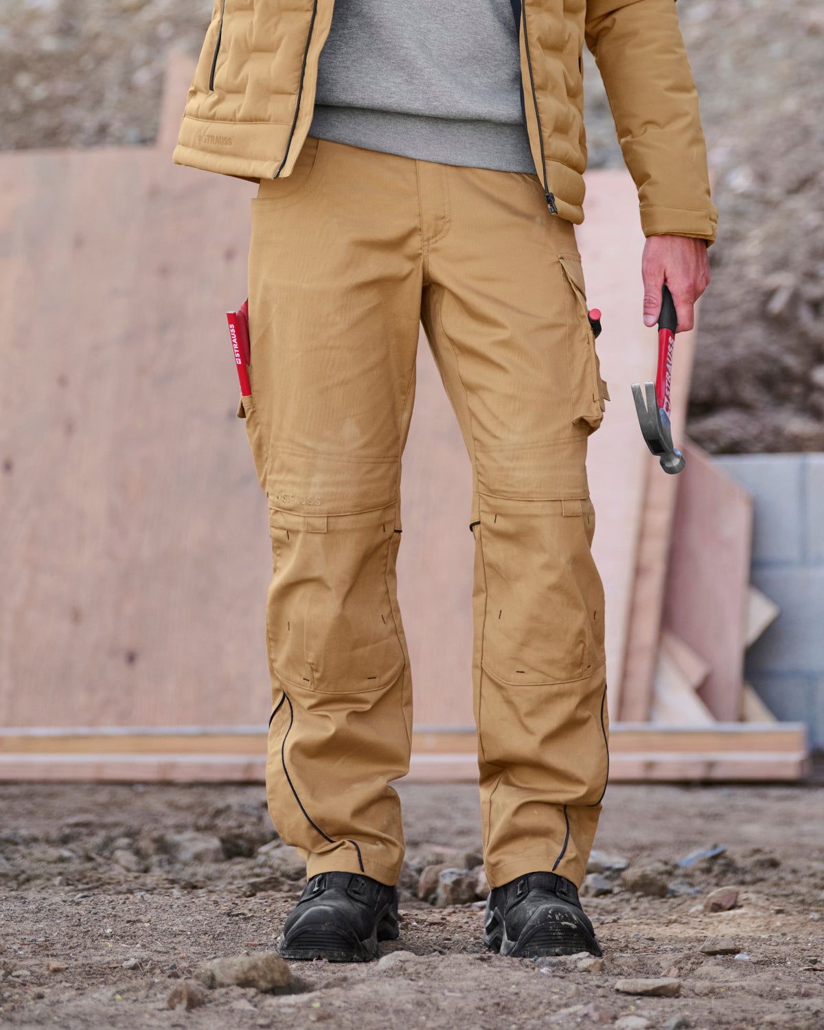 A person standing outdoors on a construction site wearing tan work pants with black utility boots, a gray sweater, and a quilted jacket, holding a hammer.