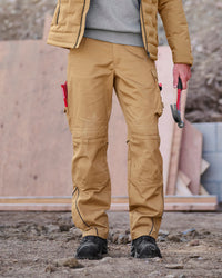 A person standing outdoors on a construction site wearing tan work pants with black utility boots, a gray sweater, and a quilted jacket, holding a hammer.