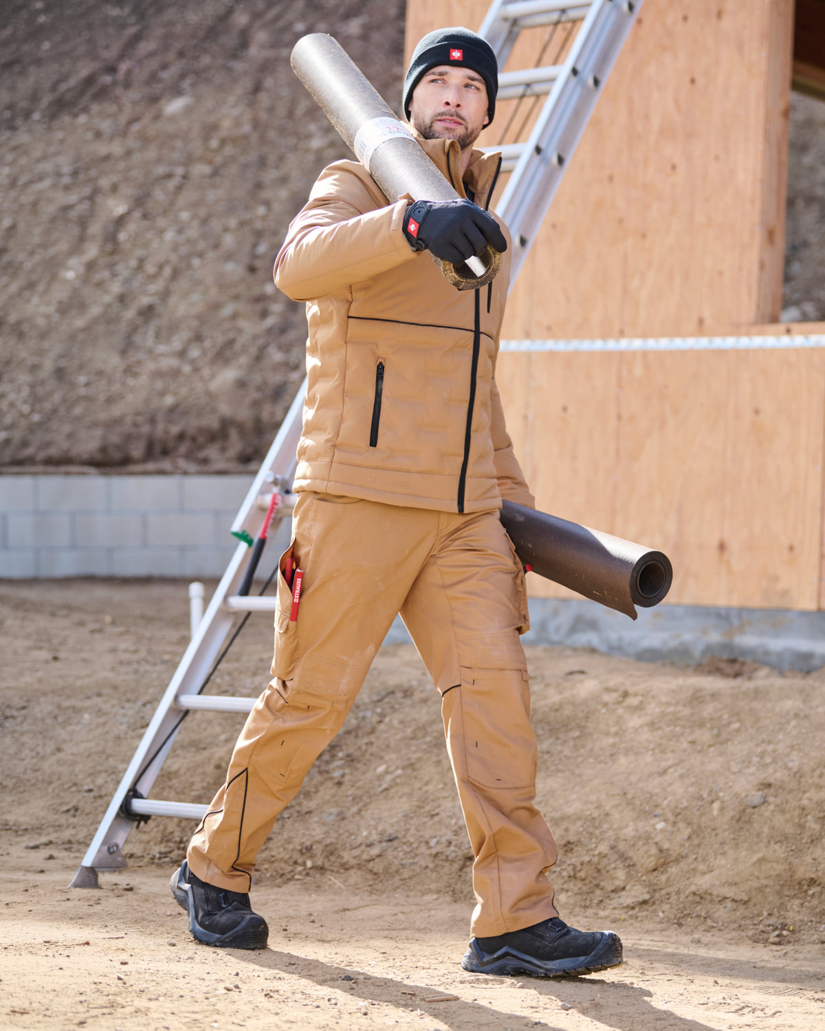 A man wearing a tan workwear jacket and matching pants stands on a construction site holding a rolled material over his shoulder, with a ladder and wooden wall in the background.