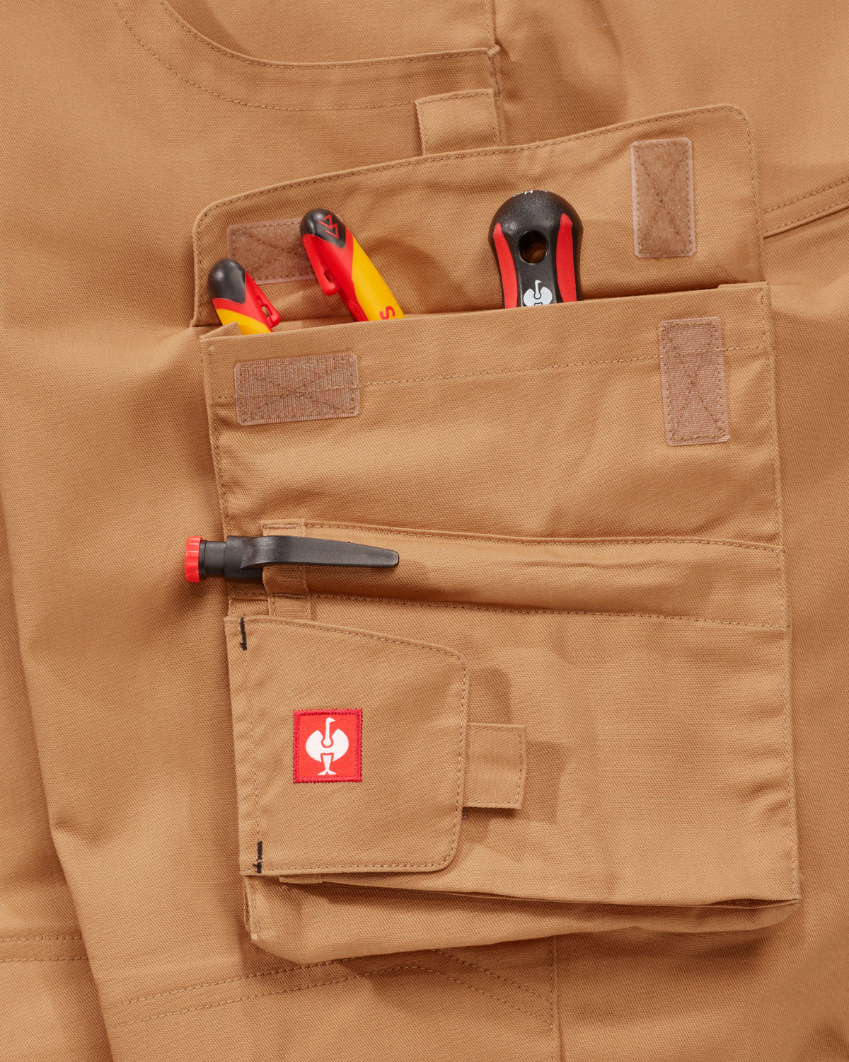 Close-up of a tan utility pocket on a matching tan garment, holding tools and a pen, with a small red STRAUSS logo patch on the pocket flap.