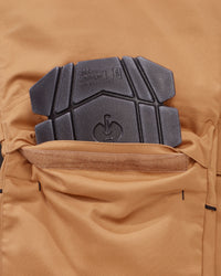 Close-up of a tan technical pants pocket with a black protective knee pad  inserted, showing fabric texture and branding embossed on the pad.