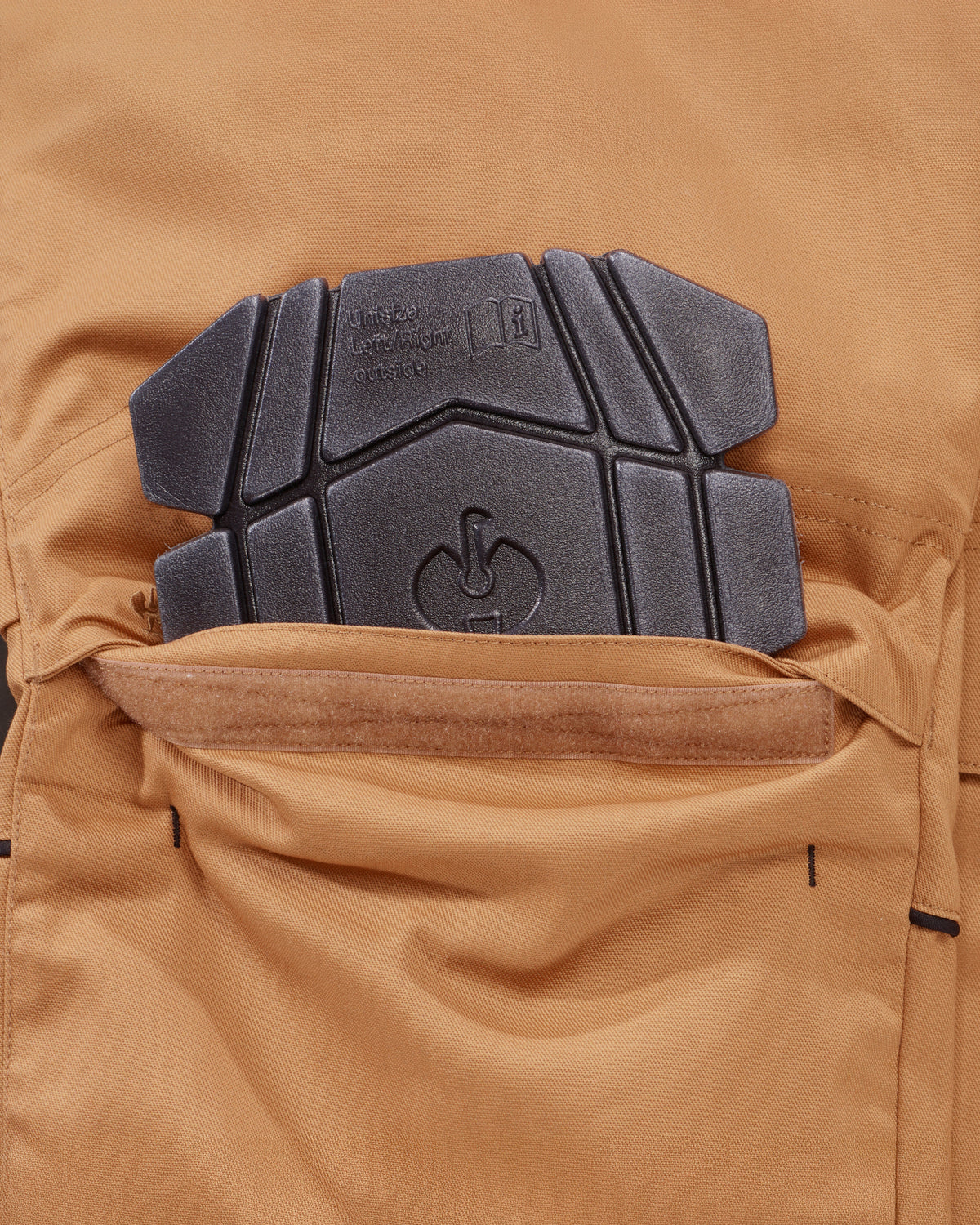 Close-up of a tan technical pants pocket with a black protective knee pad  inserted, showing fabric texture and branding embossed on the pad.