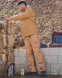 A man wearing a tan waterproof work jacket and matching cargo pants stands outdoors on a construction site next to a stone column, with tools and a black utility case nearby.